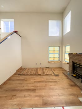 Broomfield, Colorado, USA-January 26, 2025-A spacious living room undergoing flooring installation, featuring wood-look planks partially laid out. The room has high ceilings, large windows covered with protective film, and a fireplace on the right side. Tools and construction materials indicate an ongoing home renovation project.