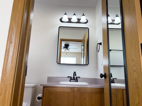 Broomfield, Colorado, USA-January 26, 2025-A newly renovated bathroom featuring a stylish vanity with wooden cabinetry, a sleek mirror, and modern light fixtures. The space is well-lit, showcasing a contemporary and functional design. A mirrored door reflects the room, adding depth to the interior.