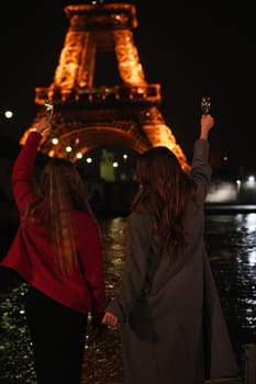 Two friends joyfully raise their glasses in front of the illuminated Eiffel Tower, capturing a moment of celebration