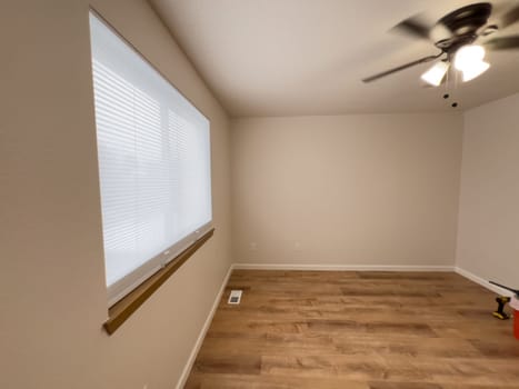 Broomfield, Colorado, USA-January 25, 2025-A freshly renovated bedroom featuring new wood-look flooring, neutral-colored walls, and a ceiling fan with lighting. The large window with blinds allows natural light to brighten the space. A few tools remain, indicating the final touches of the remodeling process.