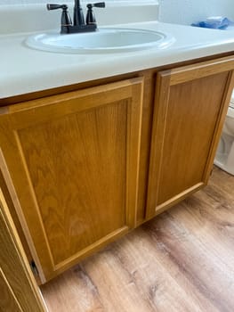 Broomfield, Colorado, USA-January 12, 2025-A close-up view of a wooden bathroom vanity featuring a white countertop, built-in sink, and black faucet. The cabinetry has a natural wood finish, complementing the newly installed wood-look flooring. The vanity adds warmth and functionality to the bathroom space.