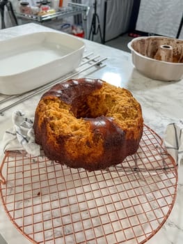 Castle Rock, Colorado, USA-December 31, 2024-A burnt bundt cake sits on a cooling rack in a food photography studio. The uneven texture and overcooked edges suggest a baking mishap. In the background, a bundt pan with cake residue and various kitchen tools are visible, highlighting the process behind the failed bake.