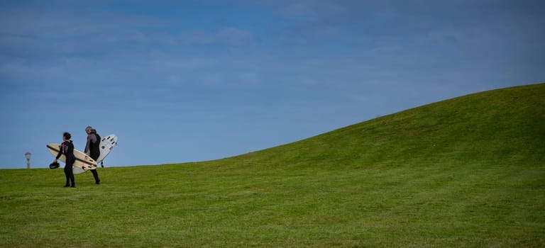Two individuals walk across a lush green hill while carrying a surfboard. The setting features a clear blue sky, suggesting a warm and sunny day perfect for surfing activities.
