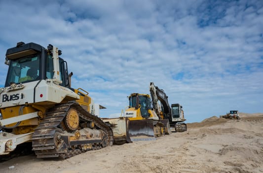 Heavy machinery is positioned along the sandy coastline, actively involved in erosion control and coastal protection efforts under a cloudy sky.