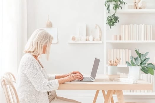 A senior woman sits at a wooden desk in a bright, airy home office, focused on her laptop. Surrounding her are plants and neatly arranged books, creating a peaceful workspace.