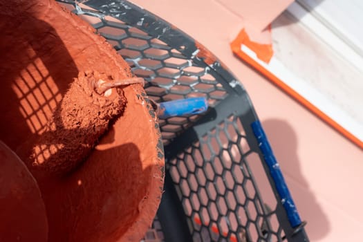 A paint roller rests in a bucket filled with reddish-brown paint. Surrounding tools and a work environment indicate ongoing home improvement activities in good weather.