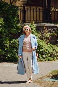 Stylish woman in blue trench coat, white trousers and coral beret walking in calm old town