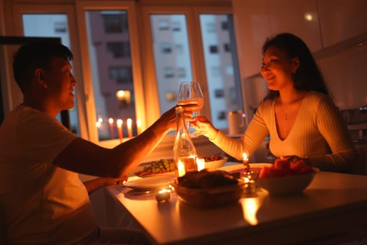 Young couple enjoying romantic candlelit dinner at home, toasting with wine glasses and celebrating their love