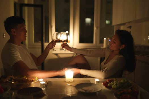 Young couple holding hands and toasting wine glasses, enjoying romantic candlelit dinner in kitchen