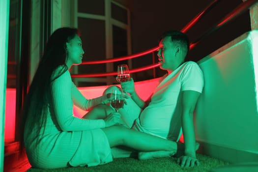 Young couple enjoying wine on their balcony at night, illuminated by vibrant neon lights, creating a romantic and modern atmosphere