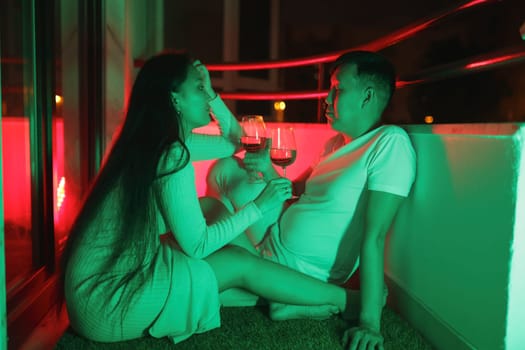 Young couple enjoying wine on their balcony at night, illuminated by vibrant neon lights