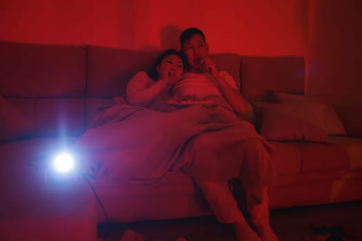 Young asian couple watching a movie projected on the wall, sitting on a sofa and eating chips