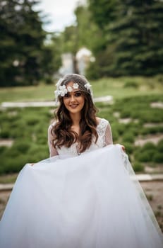 young girl bride in white dress in spring park on wedding day, beautiful brunette in long stylish dress. veil with flowers in vintage style and loose wavy hair, looks at camera