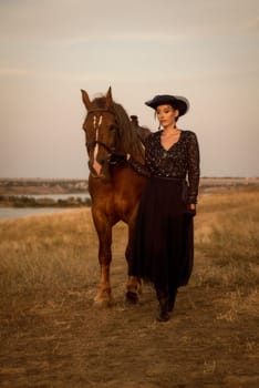 beautiful equestrian is resting after horse riding. charming girl in long dark blue dress, airy hat and gloves. leads her horse in the field in the rays of the setting sun. concept of women riding horses