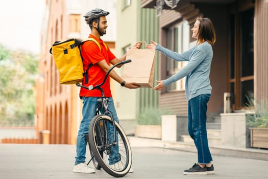 Courier with bicycle delivered no name shopping bag to client. Smiling girl takes paper bag from young man in helmet with backpack, side view, free space