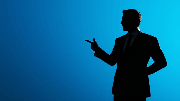 Silhouette of side view businessman in suit stands points his finger to the side at an empty space for advertising text against a blue studio background, copy space