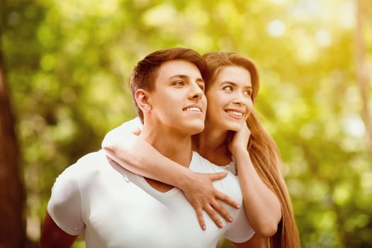 Attractive millennial guy carrying girlfriend on his back at green summer park