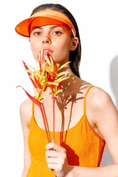 A young model in an orange sleeveless top, wearing a bright visor, holds tropical flowers, conveying summer energy and freshness, perfect for fashion and beauty imagery.