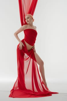 A nude woman wrapped in light red fabric poses gracefully, showcasing her beauty and elegance in a minimalist setting.