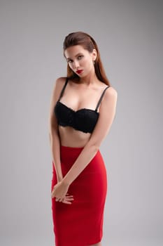 A beautiful woman stands poised in black lingerie and a red skirt, showcasing confidence in a serene studio setting.