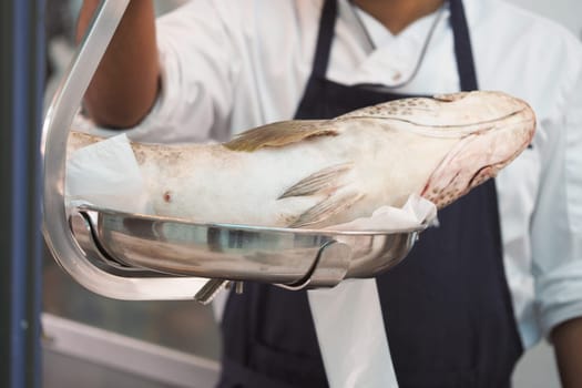 A chef presents a large fish on a stainless steel tray in a kitchen.