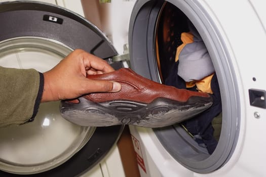 A person inserts a brown shoe into a washing machine filled with clothes.