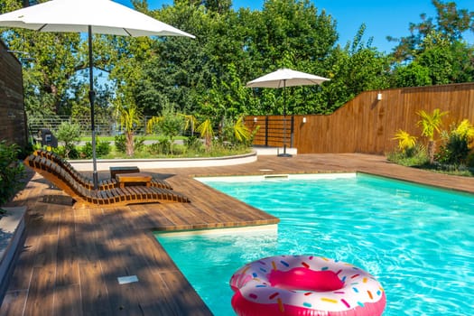 Modern backyard swimming pool with turquoise water, wooden deck, umbrellas, lounge chairs, and pink inflatable float