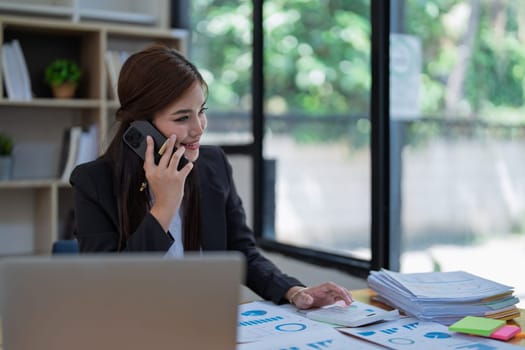 Professional businesswoman engaged in smartphone call by calculator to calculate finance with using stock marketing charts, notebook, and laptop in office.