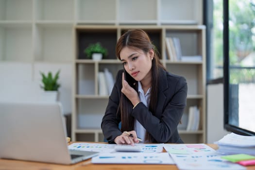 Professional businesswoman engaged in smartphone call by calculator to calculate finance with using stock marketing charts, notebook, and laptop in office.