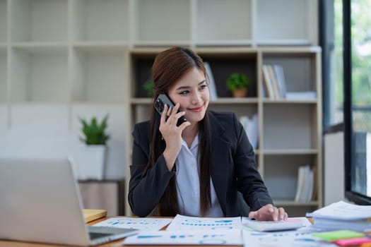 Professional businesswoman engaged in smartphone call by calculator to calculate finance with using stock marketing charts, notebook, and laptop in office.