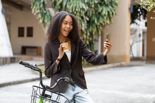 Posing with a smile, a stylish young woman holds her coffee cup and smartphone, promoting eco-consciousness as she captures the moment amidst urban greenery.