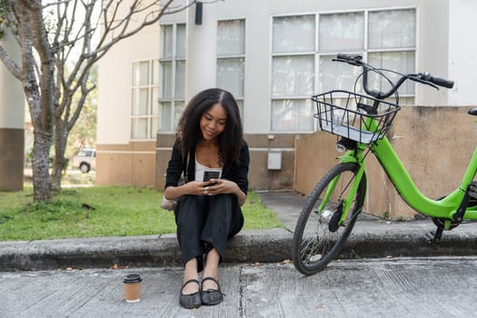 In a serene moment, the woman relaxes with her coffee and phone by her green bike, blending leisure with a sustainable urban lifestyle.