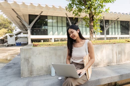 A young woman smiles while working on her laptop in a natural setting, showcasing her commitment to sustainability with her reusable cup.