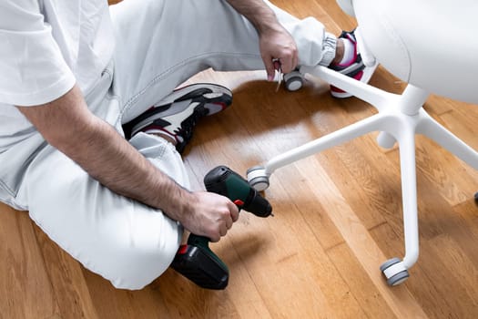 Young hispanic male assembling office chair with power drill on wooden floor.