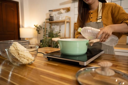 Young woman wearing apron adding water to a pot, getting ready to cook instant noodles.