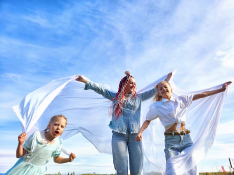 Three children are happily running and playing with white fabric in an open field beneath a clear blue sky, showcasing joy and freedom.