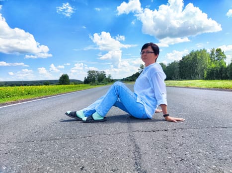 Mature blonde woman takes a relaxing break sitting on a rural road surrounded by lush greenery and blue skies.