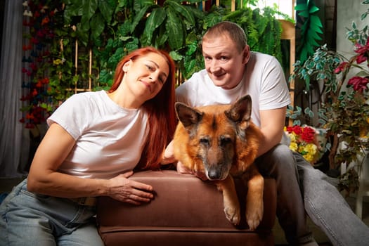A couple relaxes indoors, sitting beside their German Shepherd dog on a cozy couch surrounded by green plants.