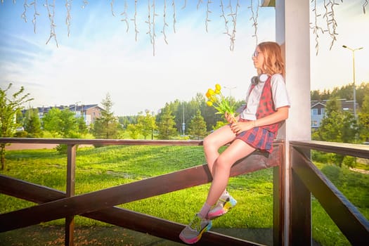 A girl with headphones enjoys a sunny day while sitting on a wooden railing, holding a bouquet of flowers in the park.
