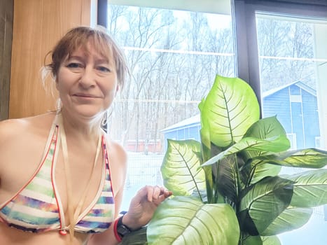 A middle-aged woman smiles while posing for a selfie next to a vibrant indoor plant, enjoying a bright and sunny moment indoors.
