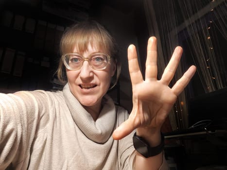 A middle-aged woman takes a fun selfie, showcasing her playful personality and cozy indoor environment at dusk.