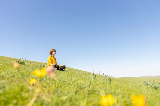 A young person sits peacefully on a green hillside surrounded by blooming wildflowers. Tranquil and dreamy spring atmosphere. Peace and connection with nature.