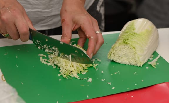 Skilled hands chop crisp cabbage on a vibrant green board, surrounded by the lively atmosphere of kitchen activity.