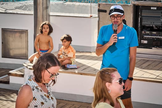Catania. Italy - March 13, 2025: Energetic entertainer in a blue shirt and white cap engaging children in a dynamic outdoor activity on a wooden deck with smiles and joy.