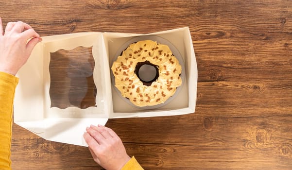 Flat lay. Carefully placing the gingerbread bundt cake, adorned with salted caramel frosting and gingerbread sprinkles, into a white paper bundt cake box with a clear window for gifting.
