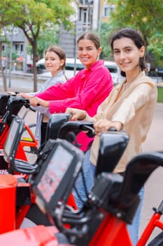 Happy young women renting bicycles in an urban park, smiling and preparing for a fun ride. Dressed casually, they enjoy a lively outdoor setting surrounded by trees and modern city buildings.