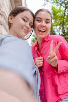 Smiling young women taking a selfie outdoors, smiling and making playful hand gestures while posing for the camera, dressed casually, capturing a fun moment in a bright and lively setting.Vertical