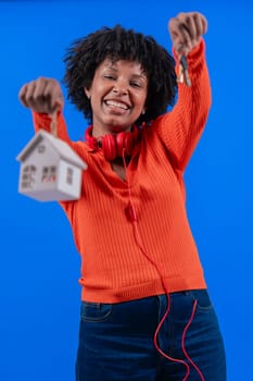Smiling African American woman holding house keys and home model against blue background.Real estate, homeownership, nvestment, success,and financial independence.Modern lifestyle and property concept