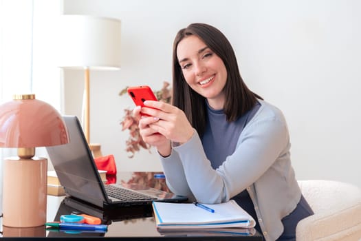 Smiling Caucasian woman holding smartphone and looking at camera. Sitting at home office with laptop, notebook, and decor. Modern workspace, technology, communication, education, and productivity.