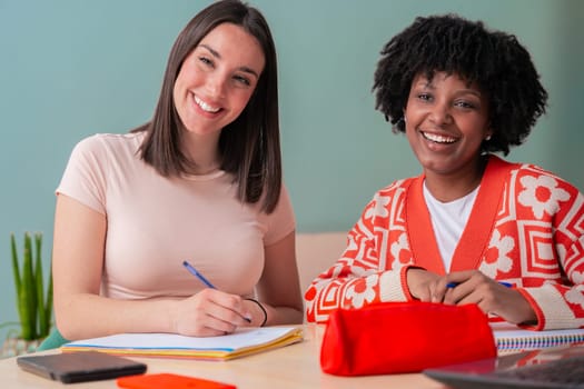 Smiling African American and Caucasian students sitting at a desk, writing in notebooks.Studying together in a modern workspace with stationery.Education, learning, collaboration, and academic success